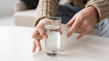 Vista parcial de un anciano con enfermedad de Parkinson y manos temblorosas tomando un vaso de agua de la mesa en casa
