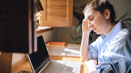 Una joven cansada ha cerrado los ojos y está sentada frente a una computadora sufriendo migraña.