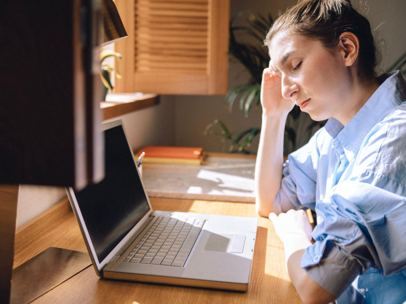 Una joven cansada ha cerrado los ojos y está sentada frente a una computadora sufriendo migraña.