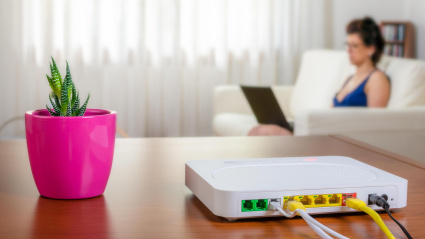 Un router sobre la mesa de una casa con una mujer al fondo usando su ordenador