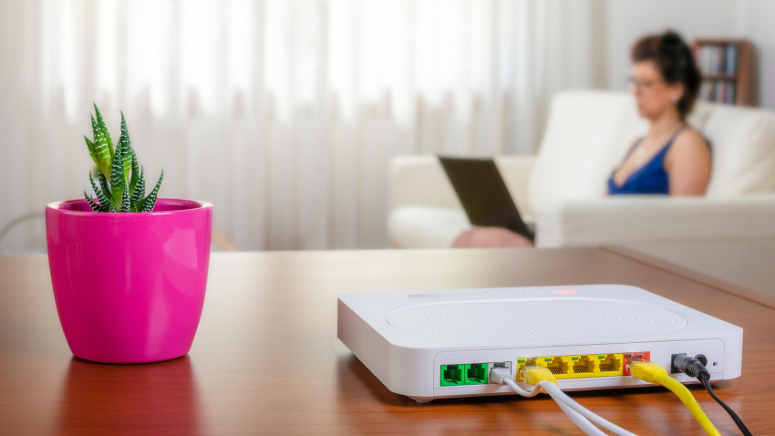 Un router sobre la mesa de una casa con una mujer al fondo usando su ordenador