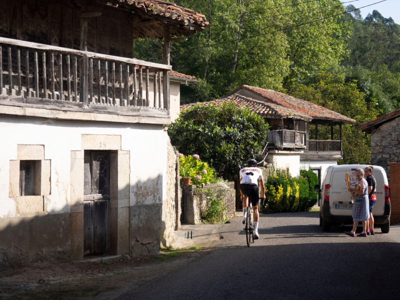 Ciclismo de carretera en Asturias, norte de España. Un ciclista de carreras pasa a dos mujeres que compran pan en una panadería móvil en el pueblo de Riocaliente en el Pico