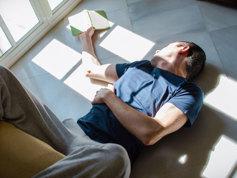 Un joven aburrido se queda en casa. Un hombre tumbado en el suelo leyendo y disfrutando del sol junto a la ventana.