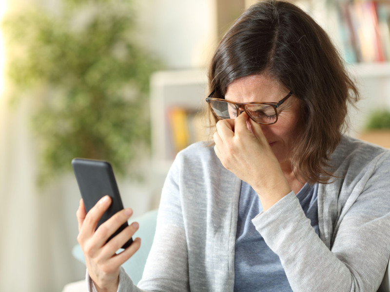 Una mujer adulta sufre fatiga visual al usar anteojos y sostener el teléfono sentada en un sofá en su casa