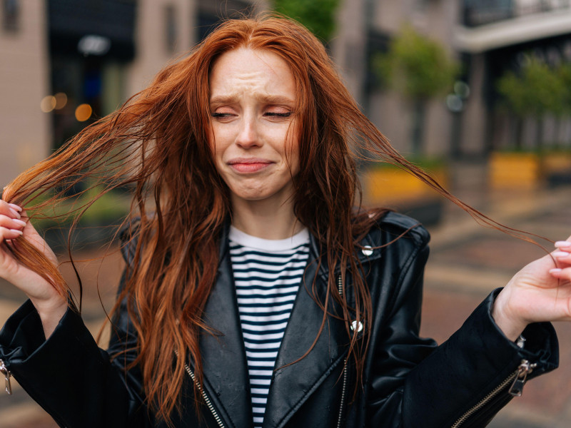 Portrait of sad attractive redhead young woman touching wet hair after autumn rain standing on beautiful city street