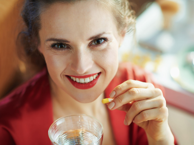 Retrato de una mujer moderna sonriente con lencería negra y bata roja tomando una pastilla con un vaso de agua en una casa moderna en un día soleado