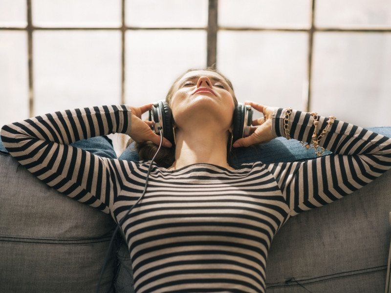 Mujer joven escuchando música con auriculares mientras está sentada en el sofá en un apartamento tipo loft