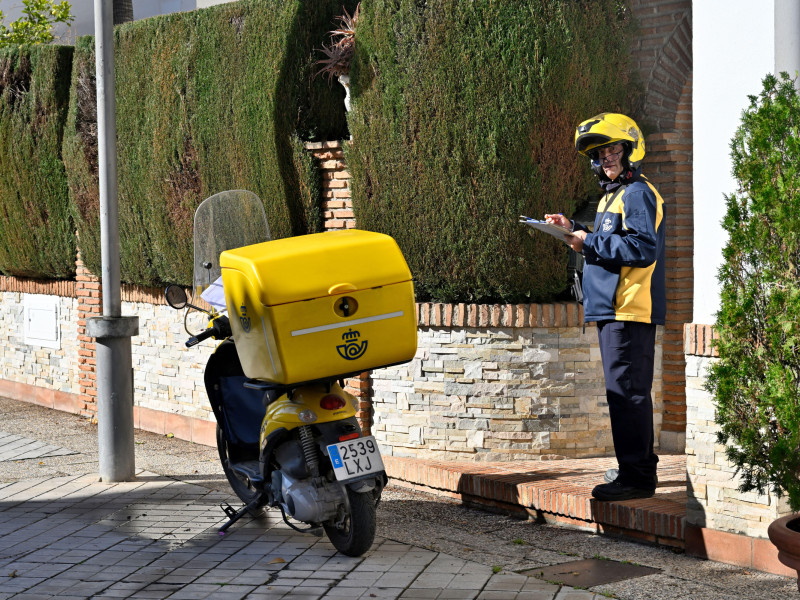 Cartero español haciendo entrega de un paquete pequeño en motocicleta en la puerta del cliente, España