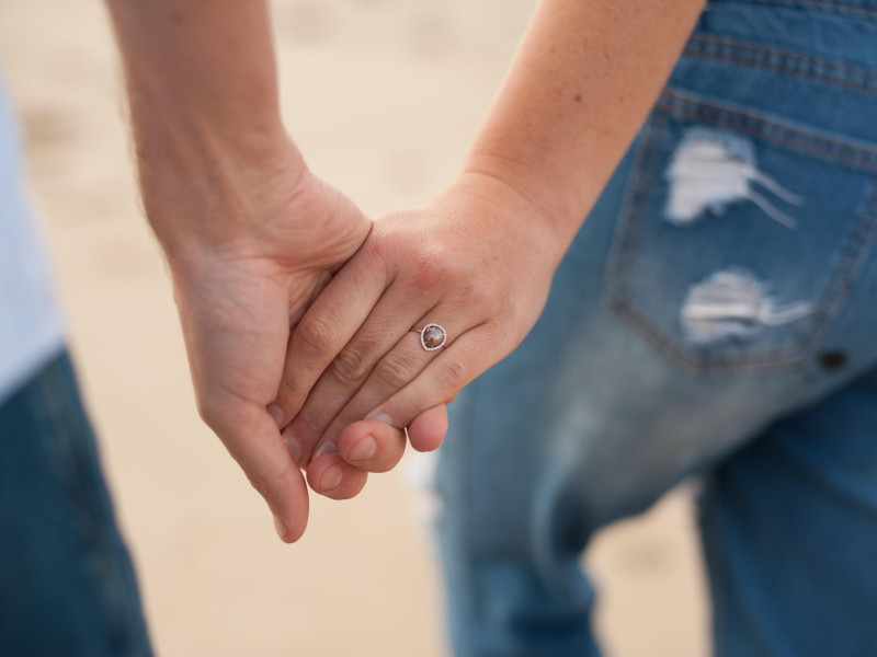 Fotografía horizontal de primer plano de una joven pareja tomada de la mano en la playa con el foco puesto en el anillo durante una sesión de fotos en la playa en un día nublado