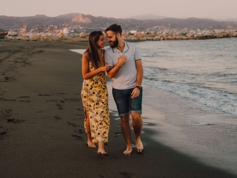 Jóvenes felices se enamoran caminando mirándose y tomándose de la mano descalzos en la playa en contraste con la luz del sol.