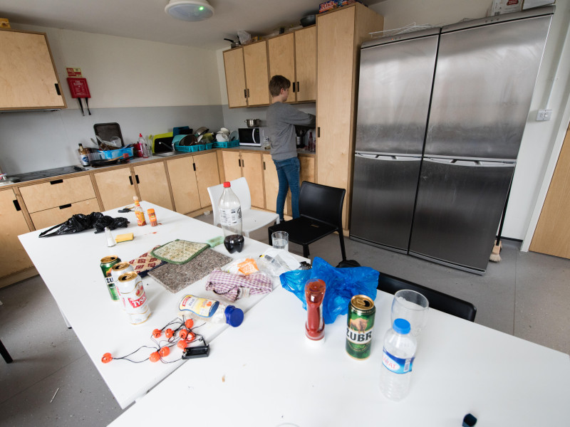 Cocina compartida de estudiantes sucia y desordenada con pilas de platos sucios junto al fregadero y una mesa común llena de basura, latas de cerveza vacías y comida.