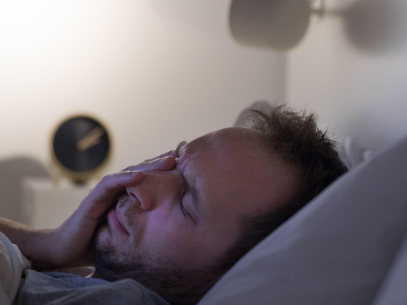 Concepto de insomnio y pesadillas. Un hombre no puede dormir después de una fiesta, se acuesta en la cama por la noche y tiene el despertador sobre la mesilla de noche. Trastornos del ciclo sueño/vigilia