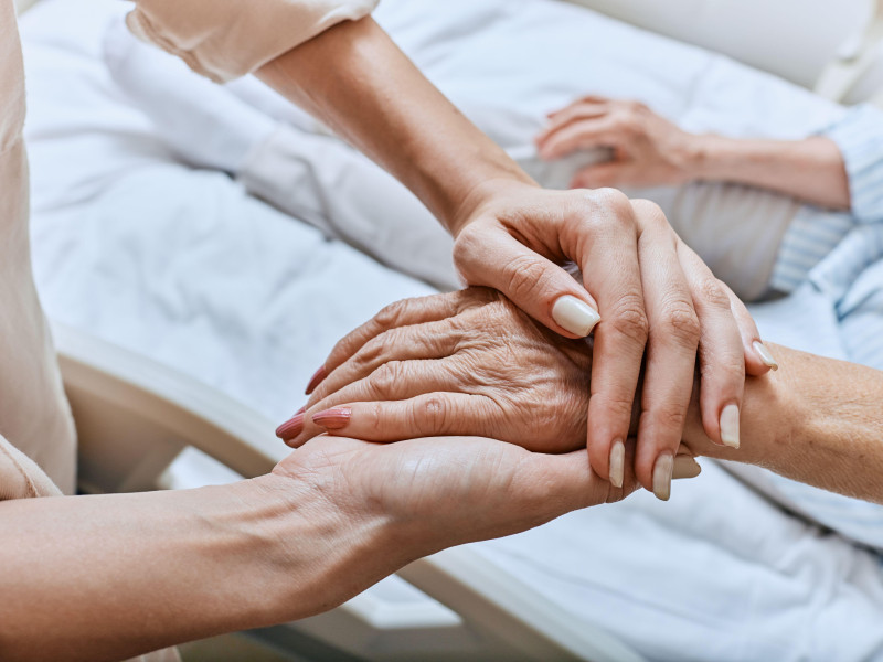 Un pariente sostiene la mano temblorosa de una mujer mayor con enfermedad de Parkinson acostada en una cama de hospital en la sala médica