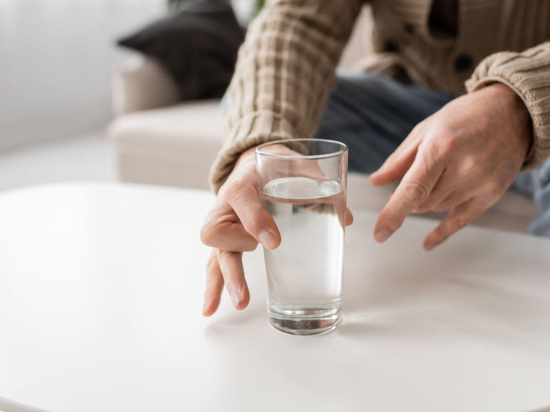 Vista parcial de un anciano con enfermedad de Parkinson y manos temblorosas tomando un vaso de agua de la mesa en casa