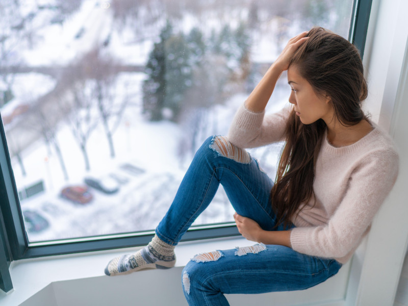 Chica triste deprimida en invierno, sola junto a la ventana de su casa mirando el clima frío, molesta e infeliz