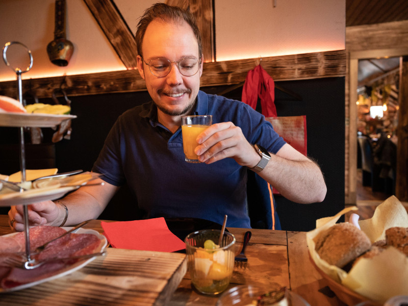 Hombre sonriente bebiendo jugo mientras está sentado en una mesa de desayuno decorada con una selección de embutidos y ensaladas saludables en primer plano