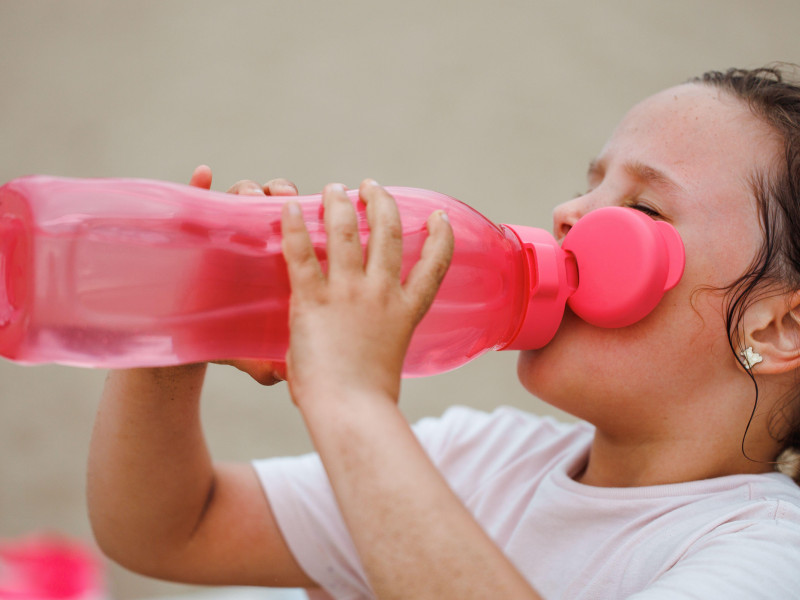 Vista lateral de una niña sedienta con cabello largo y oscuro que viste una camiseta blanca y bebe agua de una botella de plástico rosa