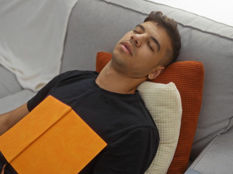 Un joven durmiendo en un cómodo sofá en una sala de estar con un libro naranja apoyado en su pecho, creando un ambiente tranquilo y relajado en el interior.