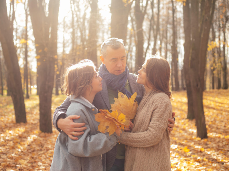 Encantadora familia de madre, padre e hija divirtiéndose juntos, abrazándose, recogiendo hojas y relajándose en bosques dorados.