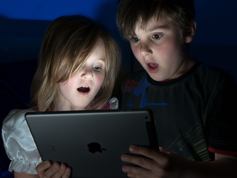 Dos niños pequeños, sorprendidos y asombrados por algo que han visto en una tableta iPad en una habitación oscura.