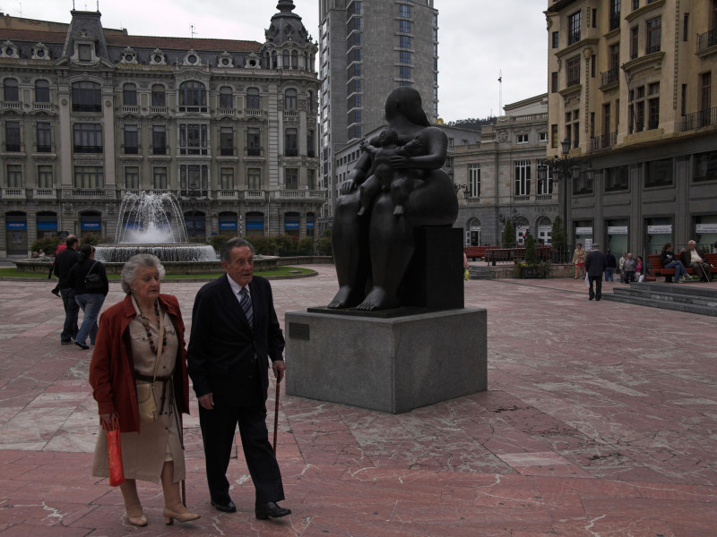 Una pareja elegantemente vestida camina por la plaza principal del distrito comercial del centro de Oviedo con una estatua de bronce de una madre amamantando a su hijo.