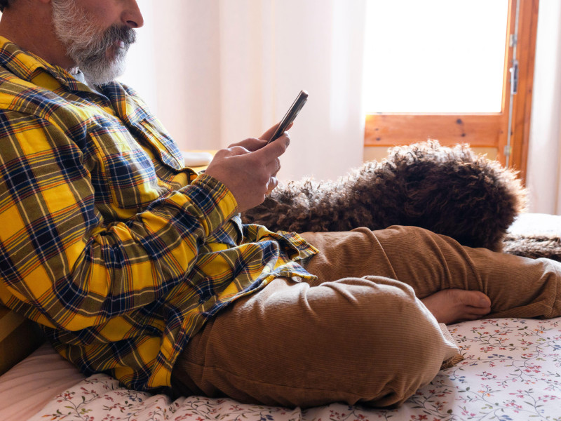 Hombre relajado usando un teléfono inteligente en el dormitorio con su perro durmiendo en la cama