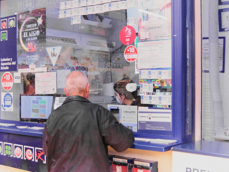 Persona mayor irreconocible frente a un puesto de lotería comprando billetes de lotería atendido por una mujer