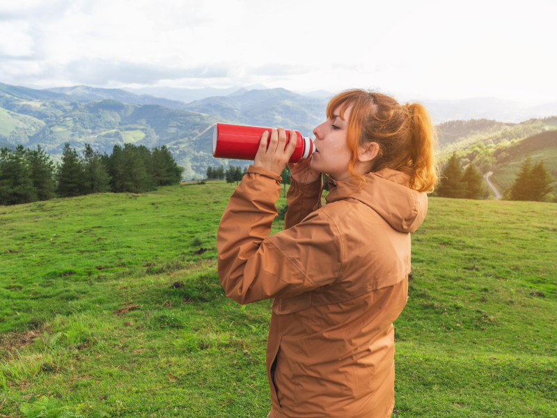Mujer joven bebiendo agua de una botella roja de pie en el campo con montañas al fondo