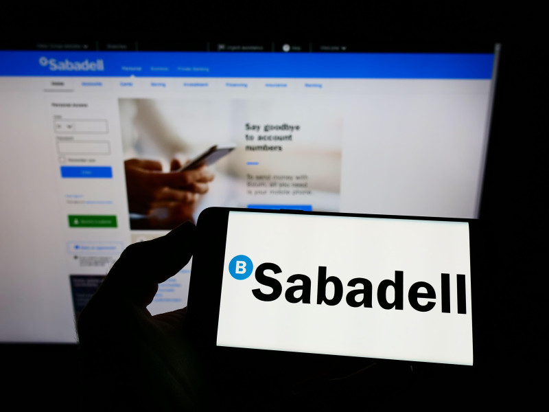 Persona sosteniendo un teléfono inteligente con el logotipo de la empresa bancaria española Banco de Sabadell S.A. en la pantalla frente a su sitio web
