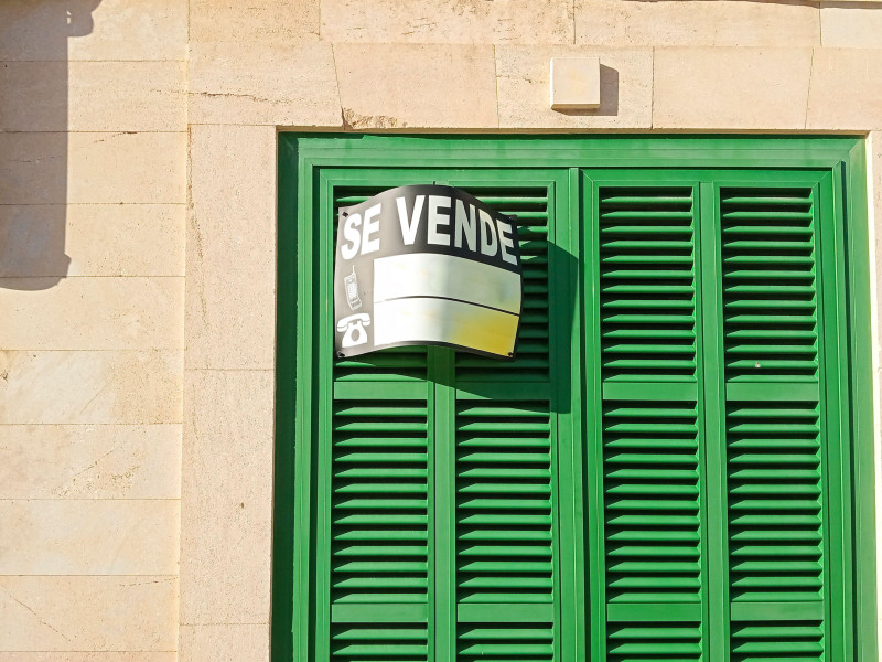 Fachada de una casa con una típica contraventana verde mallorquina, con un cartel de "se vende" escrito en español.