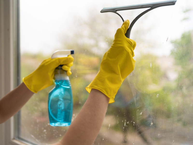Mujer con guantes amarillos lavando el cristal de la ventana con un aerosol y un trapo especiales para limpieza y desinfección.