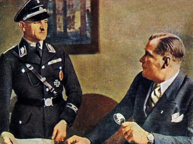 Wilhelm Bruckner (sentado a la derecha) (1884-1954) fue el ayudante principal de Adolf Hitler hasta 1940. A la izquierda, de pie, Josef "Sepp" Dietrich (1892-1966), general de las Waffen-SS alemanas y miembro del Partido Nazi de la Alemania nazi. Antes de 1929, fue chófer y guardaespaldas de Adolf Hitler, pero ascendió rápidamente tras su participación en el asesinato de los opositores políticos de Hitler durante la Noche de los Cuchillos Largos.