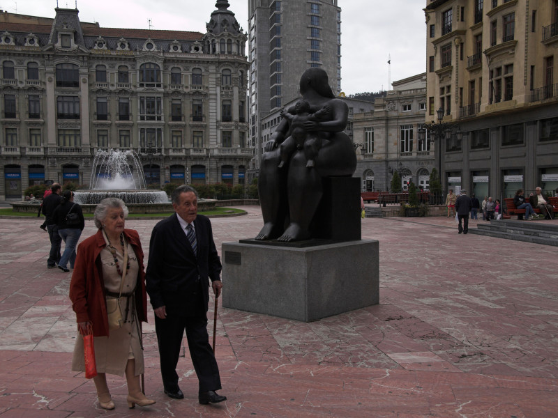 Una pareja elegantemente vestida camina por la plaza principal del distrito comercial del centro de Oviedo con una estatua de bronce de una madre lactante y su hijo.
