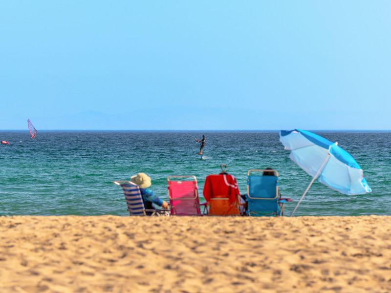 Escena de playa con gente disfrutando del sol sentada en sillas de colores con una sombrilla al lado.