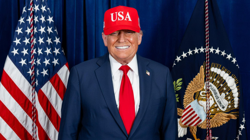 El presidente Donald Trump se encuentra frente a la bandera estadounidense y la bandera presidencial, con una gorra roja de "USA", traje oscuro, camisa blanca y corbata roja. La combinación de símbolos nacionales y ejecutivos marca el tono para un momento formal del Día de la Independencia, resaltando temas de patriotismo y autoridad. Washington