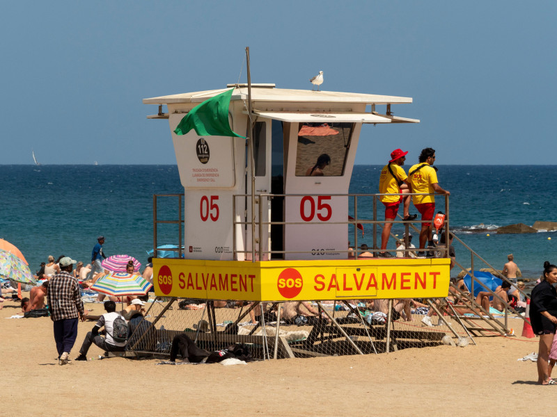 3BNKT31 Lifeguards monitor beachgoers from a rescue tower at a busy Barcelona beach, with a green flag indicating safe swimming conditions.