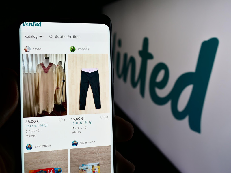Persona sosteniendo un teléfono móvil con la página web de la empresa de mercado de segunda mano en línea Vinted delante del logotipo