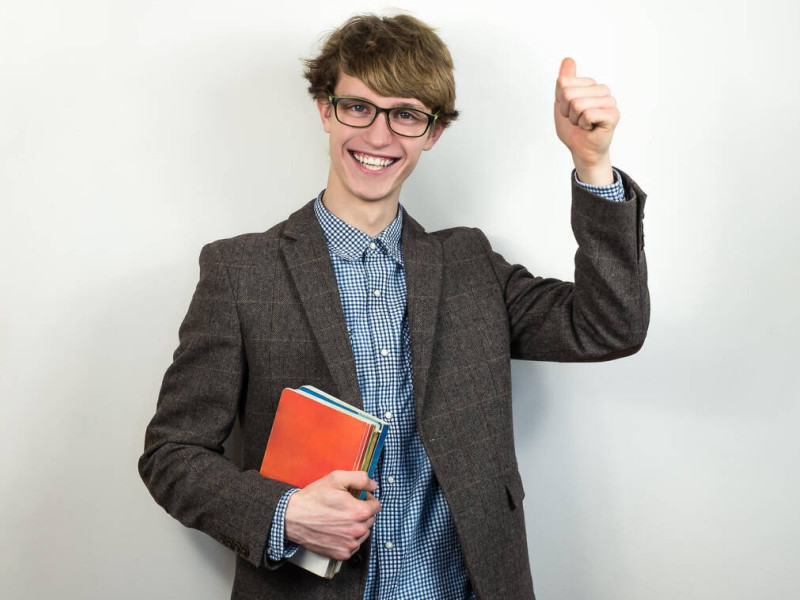 M6FRYX M6FRYX Smiling young male student makes thumbs up gesture. Handsome teenage person in tweed jacket and eyeglasses holds school books and laughs in sincere em