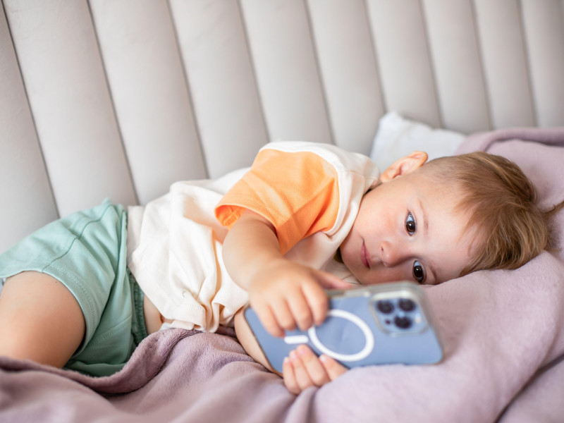 Un niño pequeño acostado mira dibujos animados en un teléfono inteligente, lo que ilustra la infancia moderna y el entretenimiento digital en el hogar.