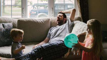 Un padre cansado bosteza en el sofá mientras sus hijos juegan a su alrededor.