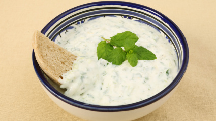 Un bol de yogur tzatziki con salsa de pepino y una rebanada de pan integral de campo sobre un mantel de mimbre.