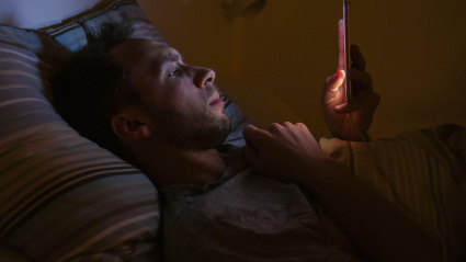 Un joven somnoliento y exhausto, acostado en la cama usando su teléfono inteligente a altas horas de la noche, no puede dormir.
