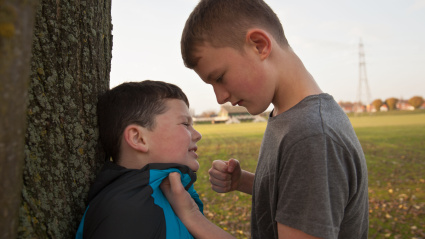 Un niño mayor acosando a otro más pequeño con el puño cerrado y sujetándolo contra un árbol.