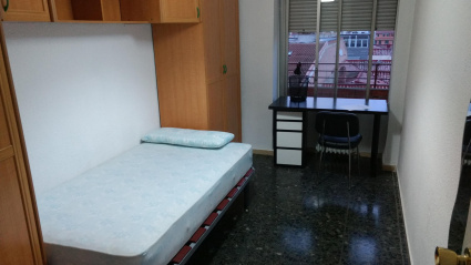 Una habitación vacía en un piso con una cama, un armario de puente, un escritorio en blanco y negro, una silla y suelo oscuro en Zaragoza.