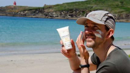Un hombre sonriente se aplica protector solar FPS 50 en la cara mientras se relaja en una playa de arena junto al mar, promoviendo la seguridad solar y unas vacaciones de verano saludables.