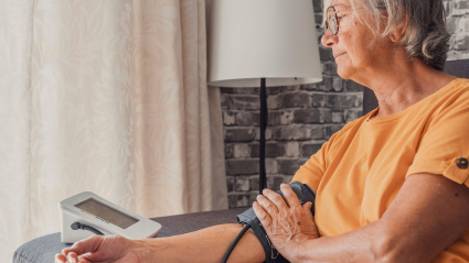 Una mujer mayor mide la presión arterial alta y baja con un medidor electrónico digital médico sentada en un sofá en casa, prevención de la aterosclerosis por enfermedades cardíacas, concepto de hipertensión e hipotensión