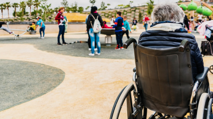 Una jubilada discapacitada sentada espalda con espalda en una silla de ruedas mientras observa a sus nietos jugar en un patio de juegos