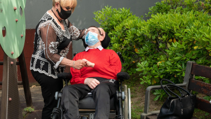 Un jubilado enfermo con una máscara en la silla de ruedas junto a una mujer que lo ayuda.