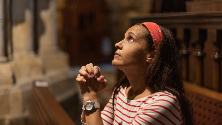 Side view of serious Hispanic female with long dark hair giving prayer with clasped hands while sitting on bench in old Catholic church and looking up