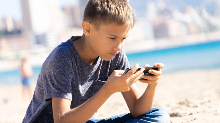 Adolescente sentado en la playa de la ciudad usando su teléfono